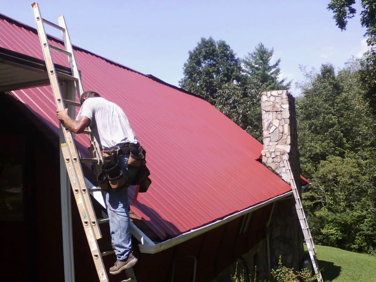 Roofer on a ladder beside a red metal roof and stone chimney — Soffit & Fascia Repair by Roofing Brothers in Mooresburg